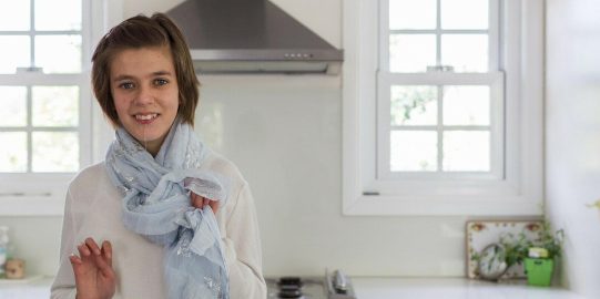 Lara, una joven blanca parada en una cocina luminosa, mirando a la cámara y sonriendo