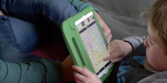 Hugo op de bank met iPad waarop Proloquo2Go te zien is