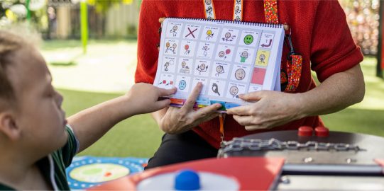 Girl pointing at symbol in PODD book while teacher holds the book