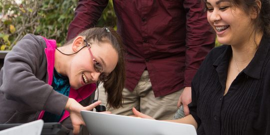 Drie mensen achter laptop, één gezicht verborgen, twee anderen glimlachen, wijzend en kijkend naar het scherm