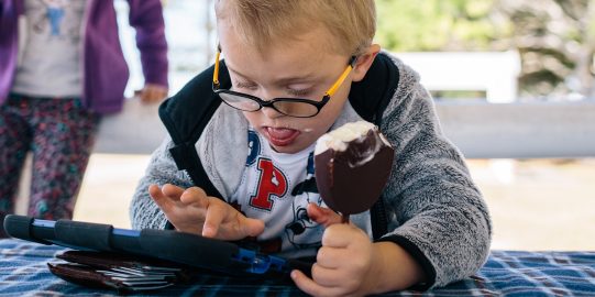 Young boy eating ice cream and using Proloquo2Go on iPad and a person behind him