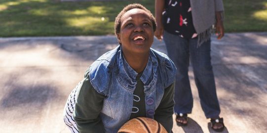 Girl looking up with basketball in her hands, ready to throw, adult in background