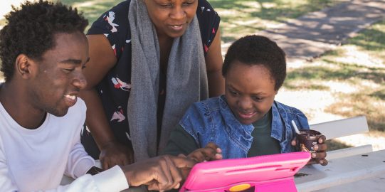 Woman, young man and child smiling and looking and pointing at iPad in a park