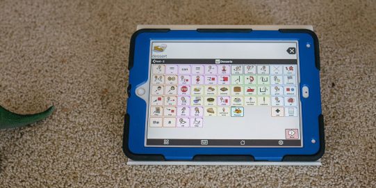 iPad met Proloquo2Go 11x7 rooster op tapijt met speeltjes ernaast.