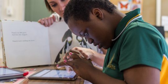 Leerlinge type op iPad terwijl lerares boek vasthoudt en toekijkt.