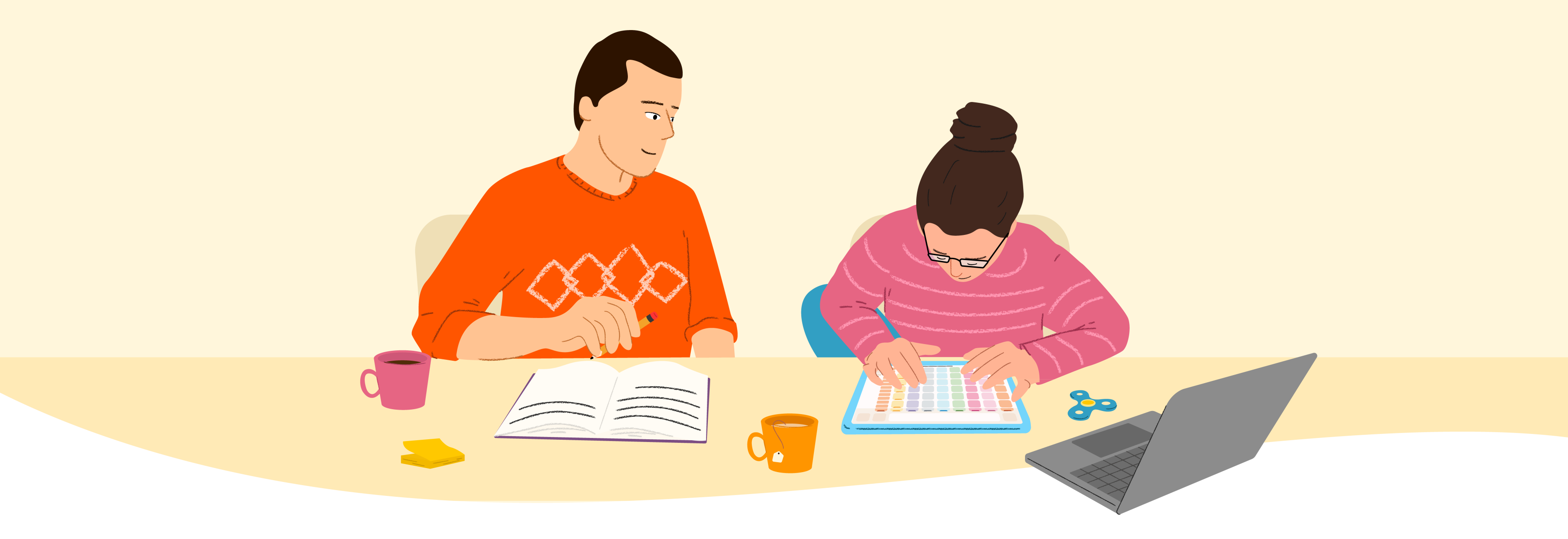 Two people at a table, one writing, one tapping on an AAC device, with a laptop, mugs, and a fidget spinner.