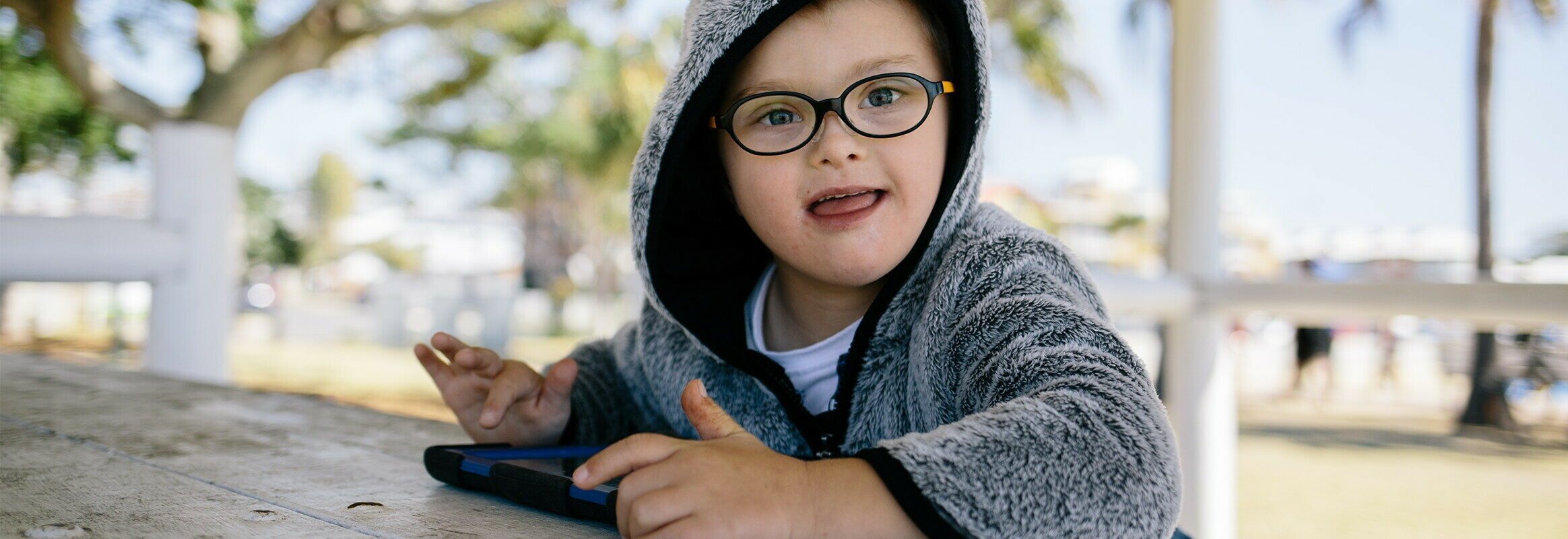 Lucas, een kleine witte jongen in een hoodie zit buiten, met een iPad en lacht