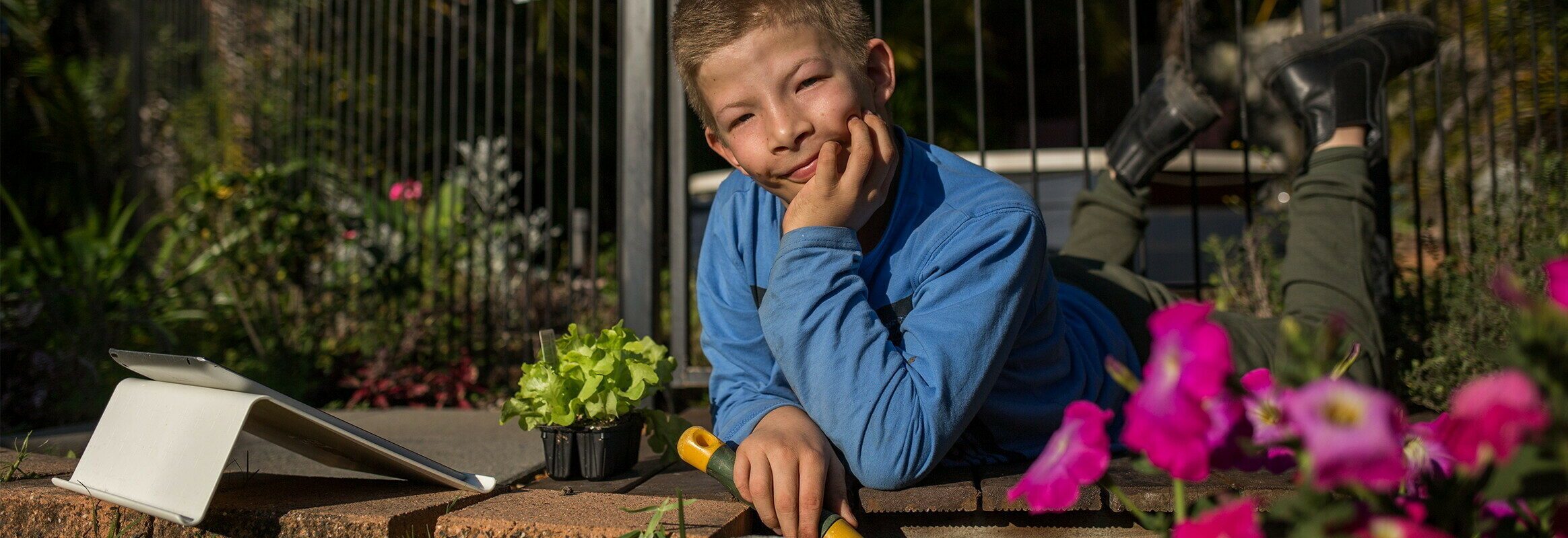 Tom, a white teenage boy laying down and working in the garden with an iPad at his side