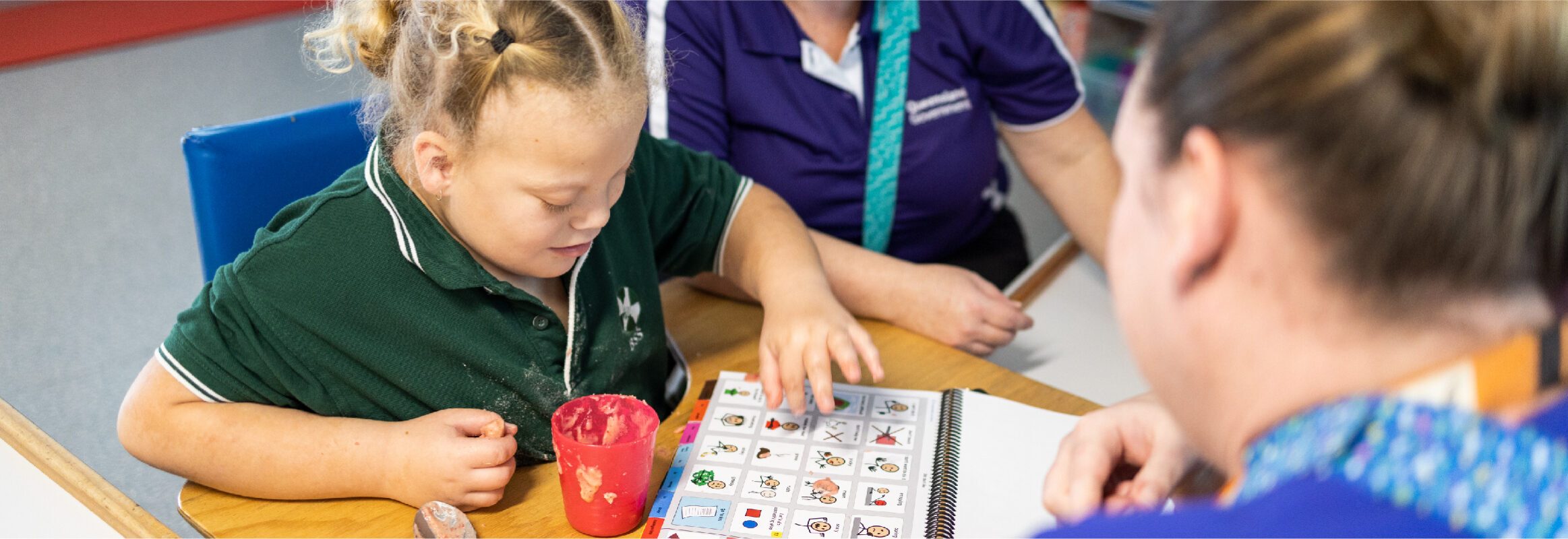 Girl in classroom with PODD print book on desk, pointing to a symbol while two people watch