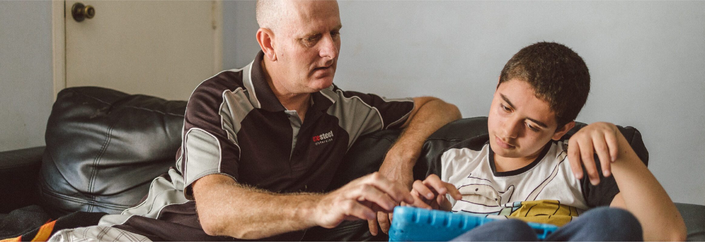 Son using iPad as communication device to talk to his father, both sitting on a sofa inside a house.