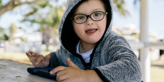 Lucas, un petit garçon blanc en sweat à capuche, assis dehors, utilisant un i Pad et souriant