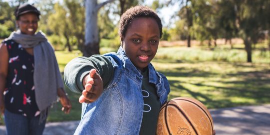 Bonita, een tienermeisje uit Malawi op het basketbalveld, gebarend naar de camera, met een andere zwarte vrouw op het veld op de achtergrond