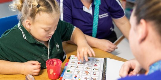 Girl in classroom with PODD print book on desk, pointing to a symbol while two people watch