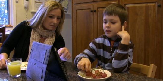 Moeder en zoon aan keukentafel, druiven etend en communiceren met iPad