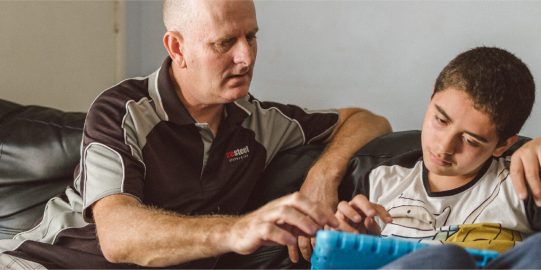 Son using iPad as communication device to talk to his father, both sitting on a sofa inside a house.