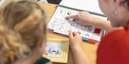 Student and teacher seen from the back with a PODD book in front of them on the table. Teacher pointing at a symbol in the PODD book.