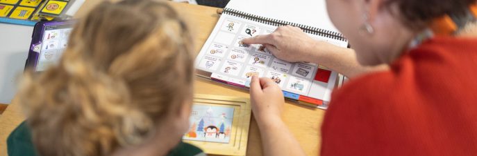 Student and teacher seen from the back with a PODD book in front of them on the table. Teacher pointing at a symbol in the PODD book.