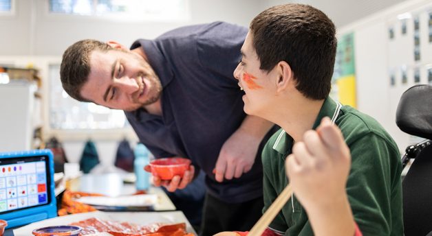 Young male student with orange paint on his face smiling at teacher who holds the cup with the paint.