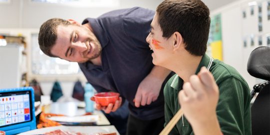 Young male student with orange paint on his face smiling at teacher who holds the cup with the paint.