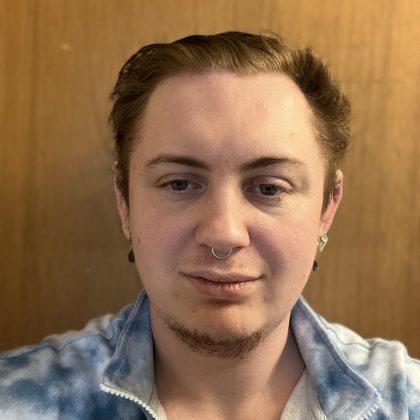 A young white masculine person smiles at the camera They have multiple facial and ear piercings and are wearing a blue and white sweatshirt